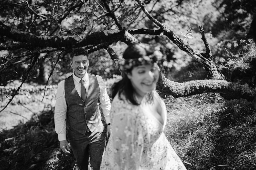 A man in a suit and a woman in a floral dress stand under a tree. The woman is in the foreground, and both are smiling. The scene is outdoors with abundant greenery.