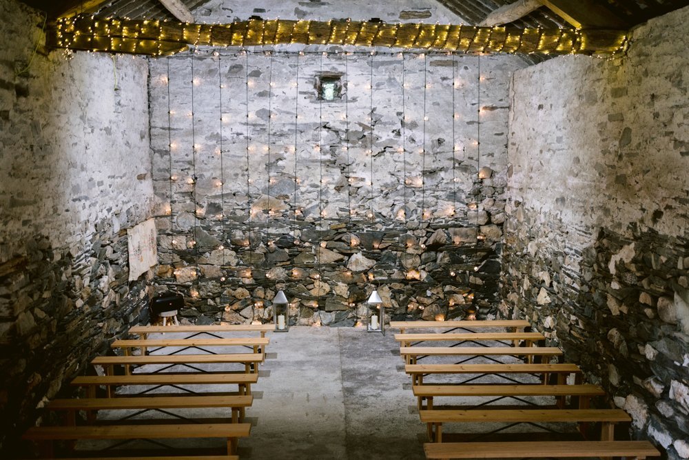 Stone-walled room with wooden benches, string lights on the ceiling and walls, lanterns on the floor, and a small window high on the far wall.