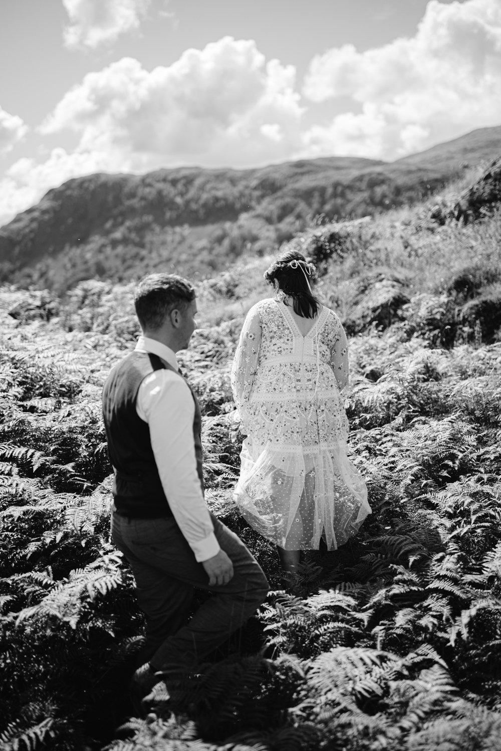 A couple in formal attire walks through a fern-covered landscape with mountains in the background under a cloudy sky. The image is in black and white.