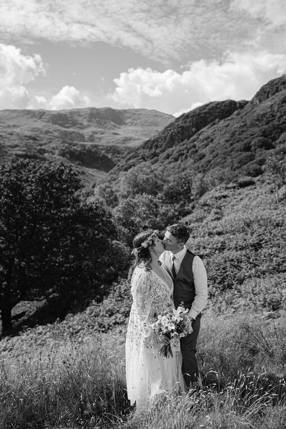 A bride and groom kiss outdoors in a mountainous landscape. The bride holds a bouquet and wears a floral crown. The scene is captured in black and white.