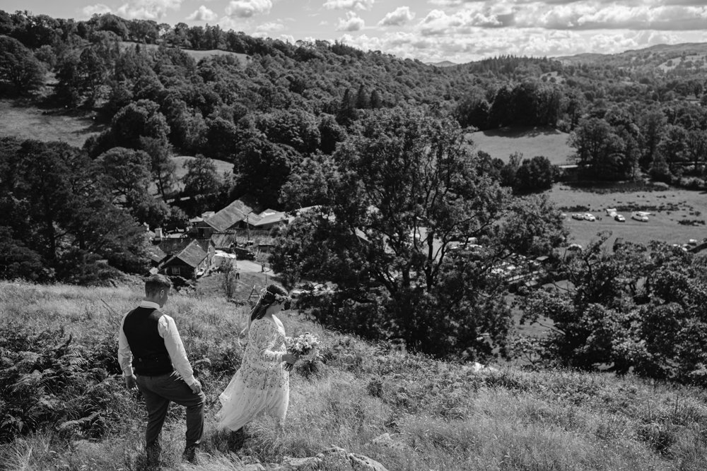 A couple in wedding attire walks down a grassy hill overlooking a rural landscape with trees and scattered buildings under a partly cloudy sky.