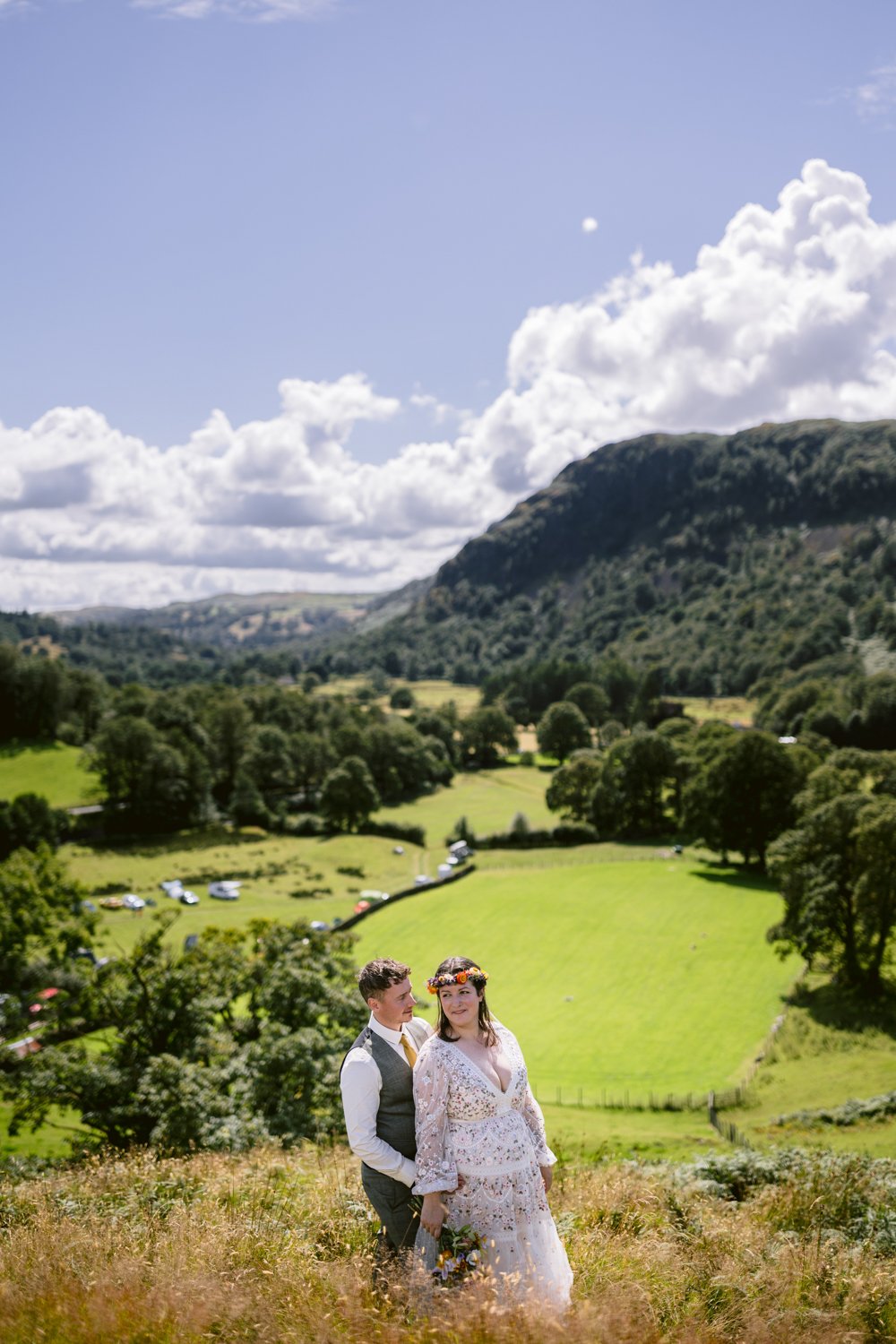 A couple dressed in wedding attire stands in a grassy field, with rolling hills and a partly cloudy sky in the background.
