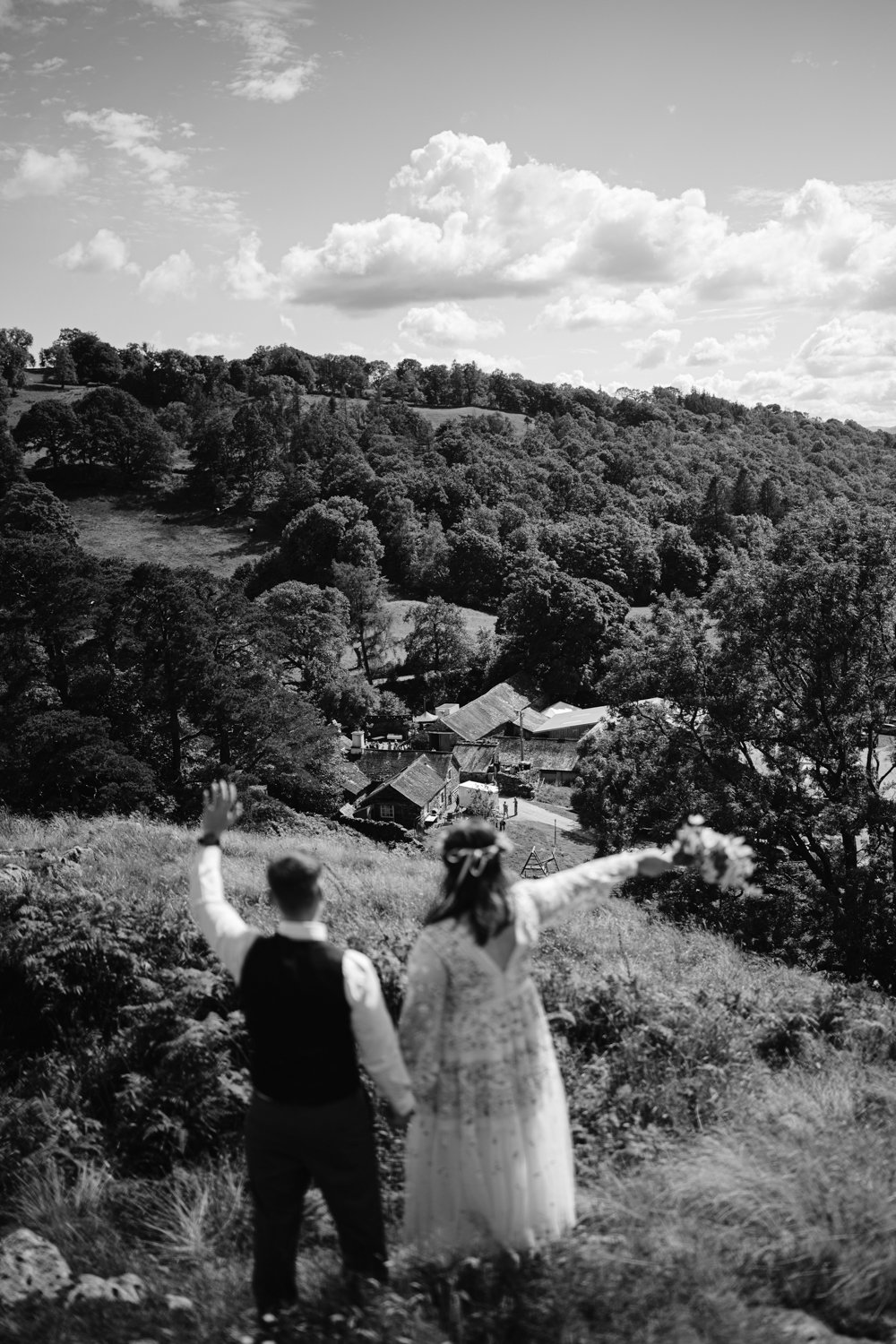 A couple in wedding attire stands on a hill, holding hands and waving towards a rural village set in a lush, tree-covered landscape under a partly cloudy sky. Black and white image.