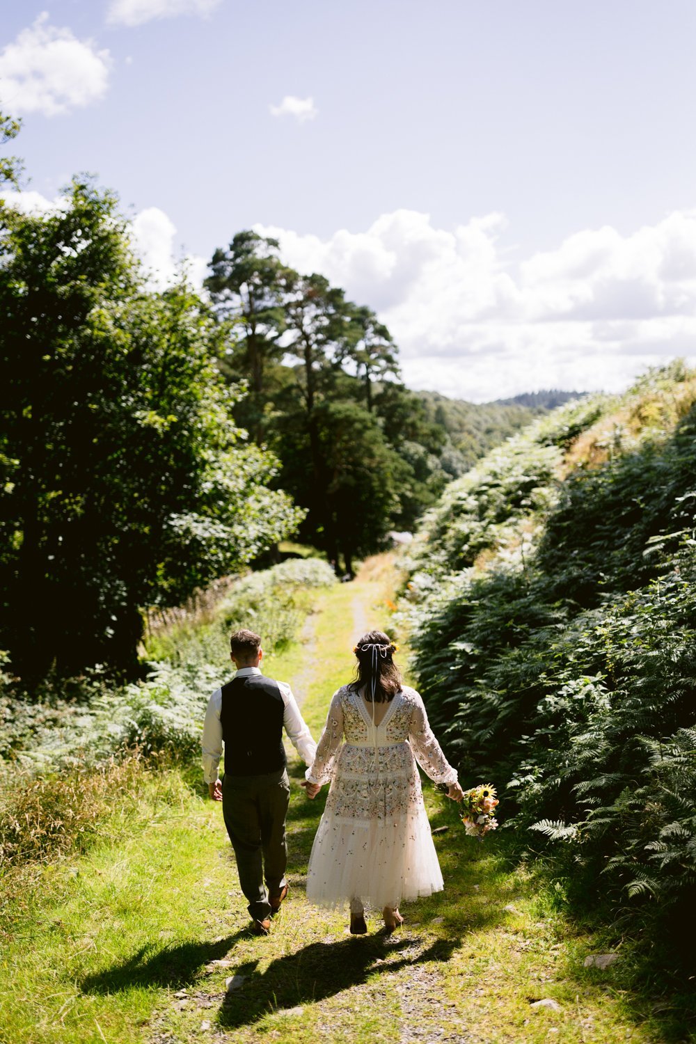 A couple in wedding attire walks hand in hand down a sunlit forest path surrounded by greenery and scattered clouds in the sky.