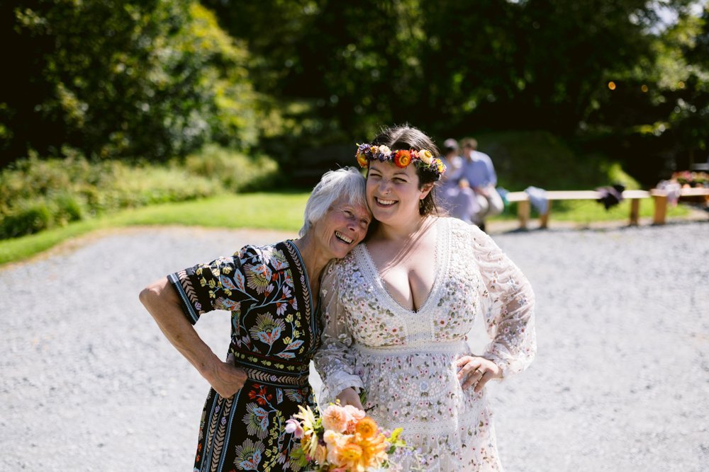 Two women outdoors, smiling. The older woman leans on the younger, who is holding a bouquet and wearing a floral crown. Both wear floral-patterned dresses.