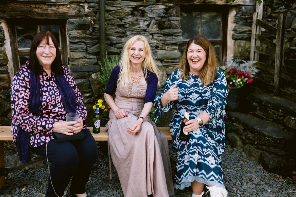 Three women sitting on a wooden bench outside, laughing and holding drinks, with stone walls in the background.