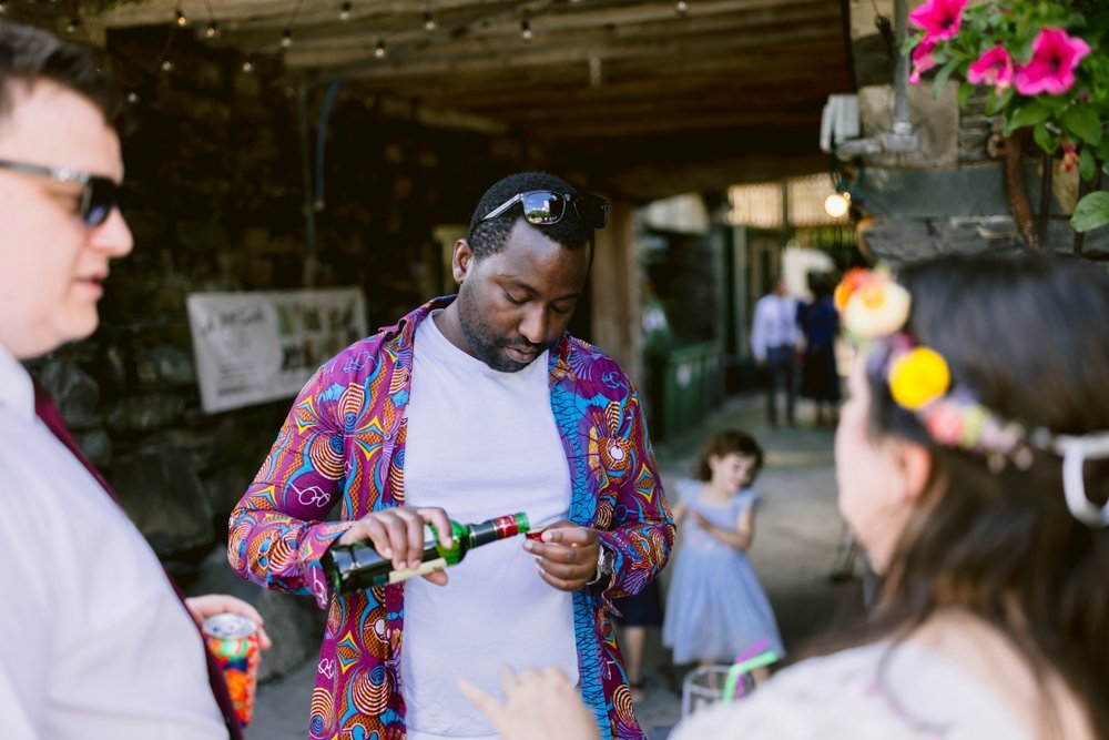 A man in a colorful shirt opens a bottle outdoors. Two people, including a person in a flower crown, are nearby. A child in the background walks toward a dark corridor.