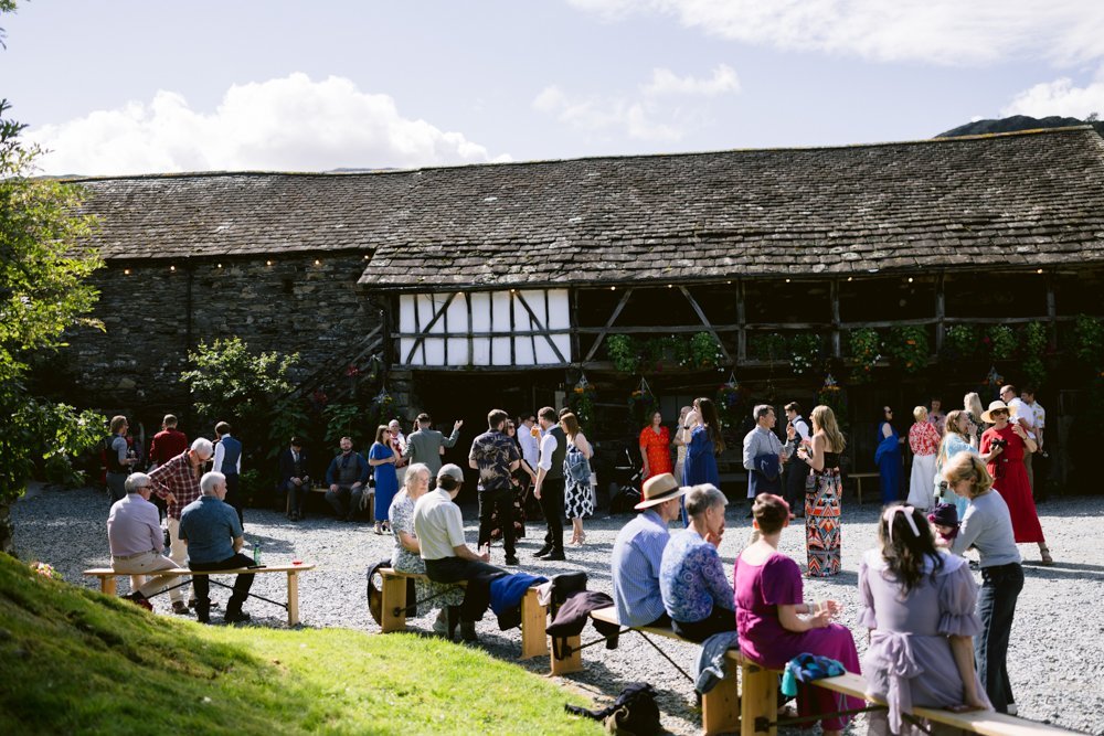 A diverse group of people gathers outdoors near a rustic barn. Some are seated on benches, while others stand and interact. The scene is bathed in sunlight.