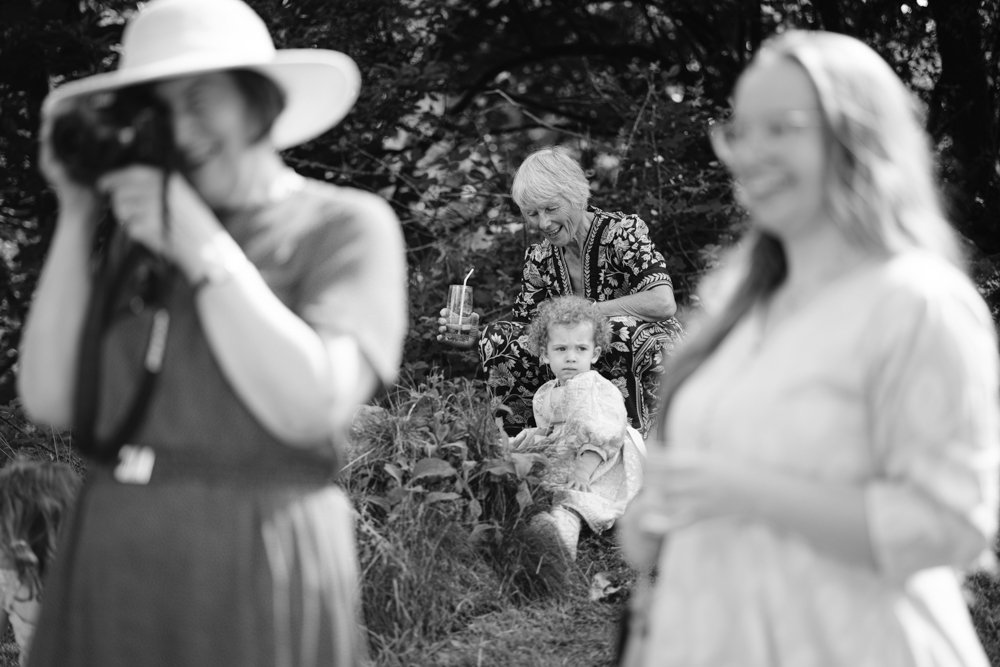 A monochrome photo shows a woman photographing, another smiling, and an older woman seated with a drink near a young girl outdoors.