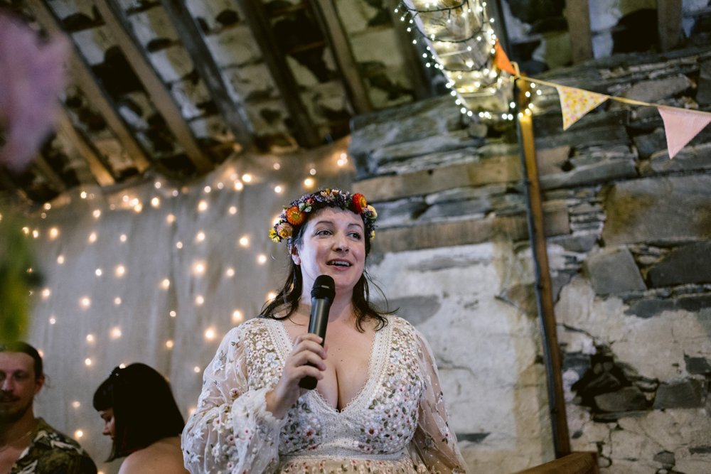 A woman in a floral dress and flower crown speaks into a microphone in a rustic setting with stone walls and string lights.