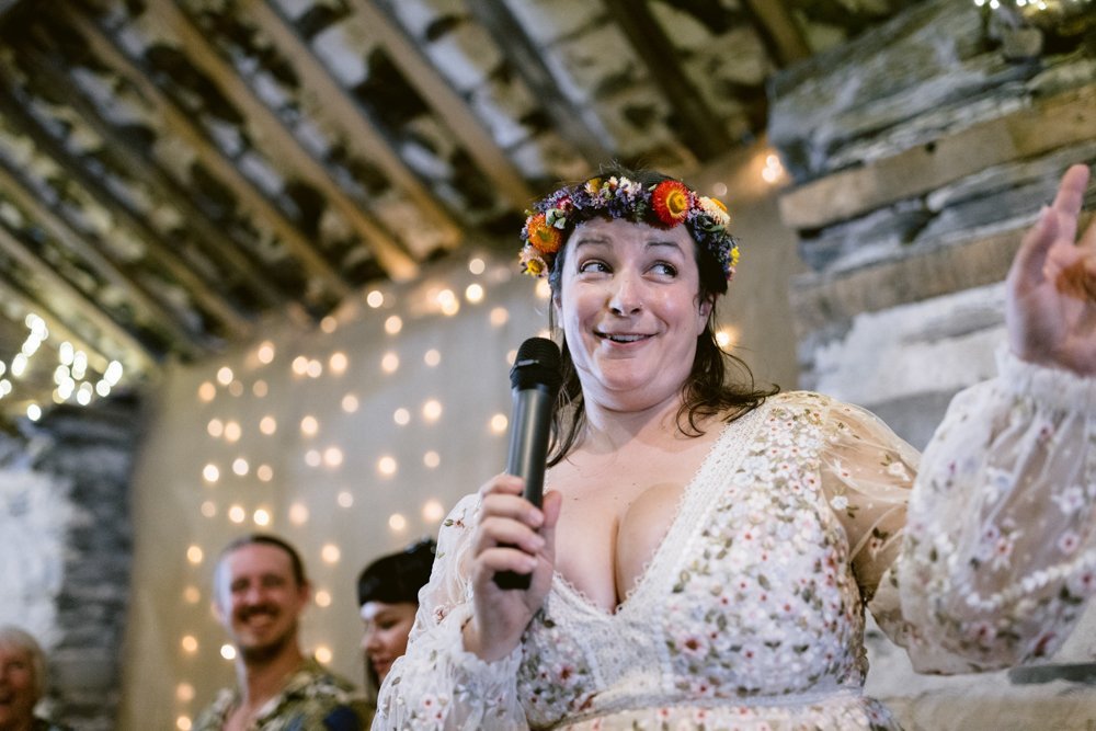A woman with a floral crown holds a microphone, gesturing while speaking. She stands indoors with string lights on the wall behind her. Two people are in the background.