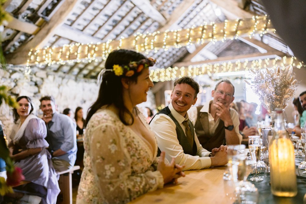 People sitting at a rustic wedding reception, with a woman in a floral crown talking. String lights hang from the wooden ceiling.
