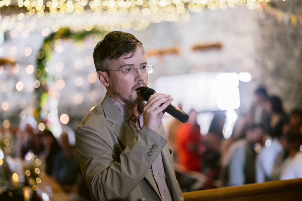 A man in a beige jacket speaks into a microphone at an indoor event with blurred lights and people in the background.