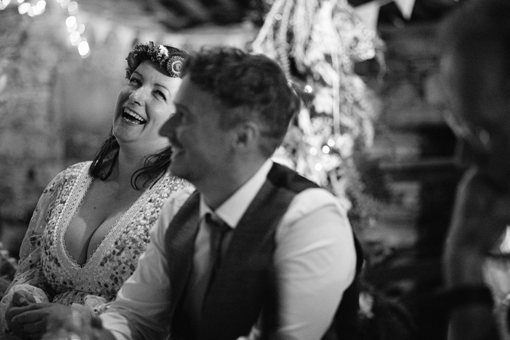 A woman in a decorated dress and a man in a vest sit together, smiling, in a festive setting with lights and bunting. Black and white photo.