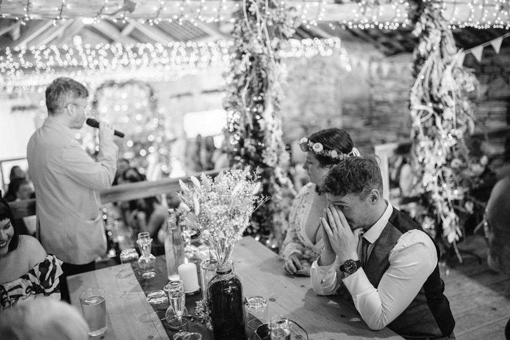 A man speaks into a microphone at a rustic indoor event. A couple listens at a table adorned with flowers and candles. The room is decorated with string lights and garlands.