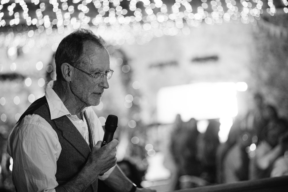 A man with glasses and a vest speaks into a microphone at an indoor event, with string lights hanging above and an audience in the background.
