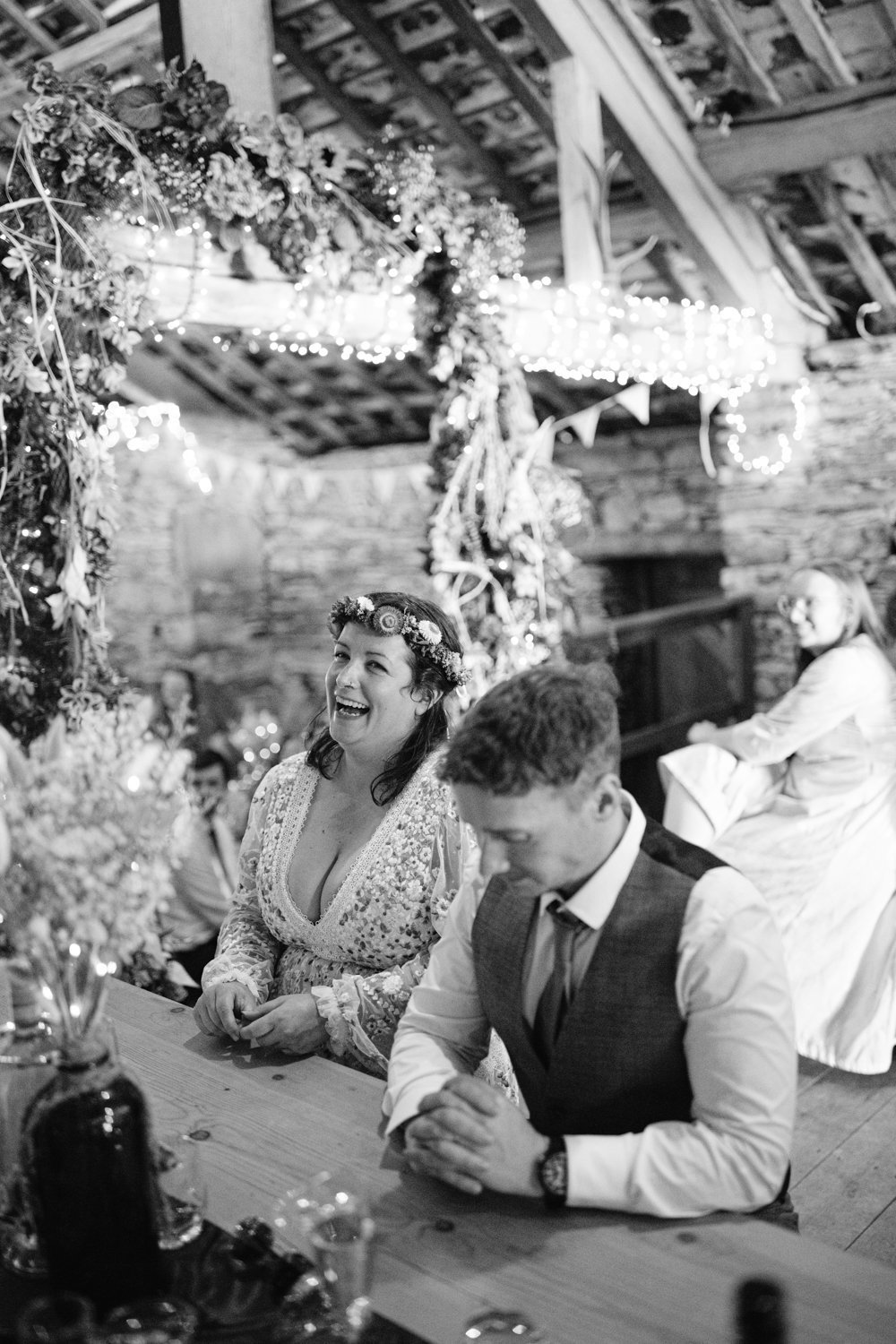A woman with a floral crown and a man in a suit sit at a decorated table in a rustic setting. Another person sits in the background. The scene has a festive atmosphere.