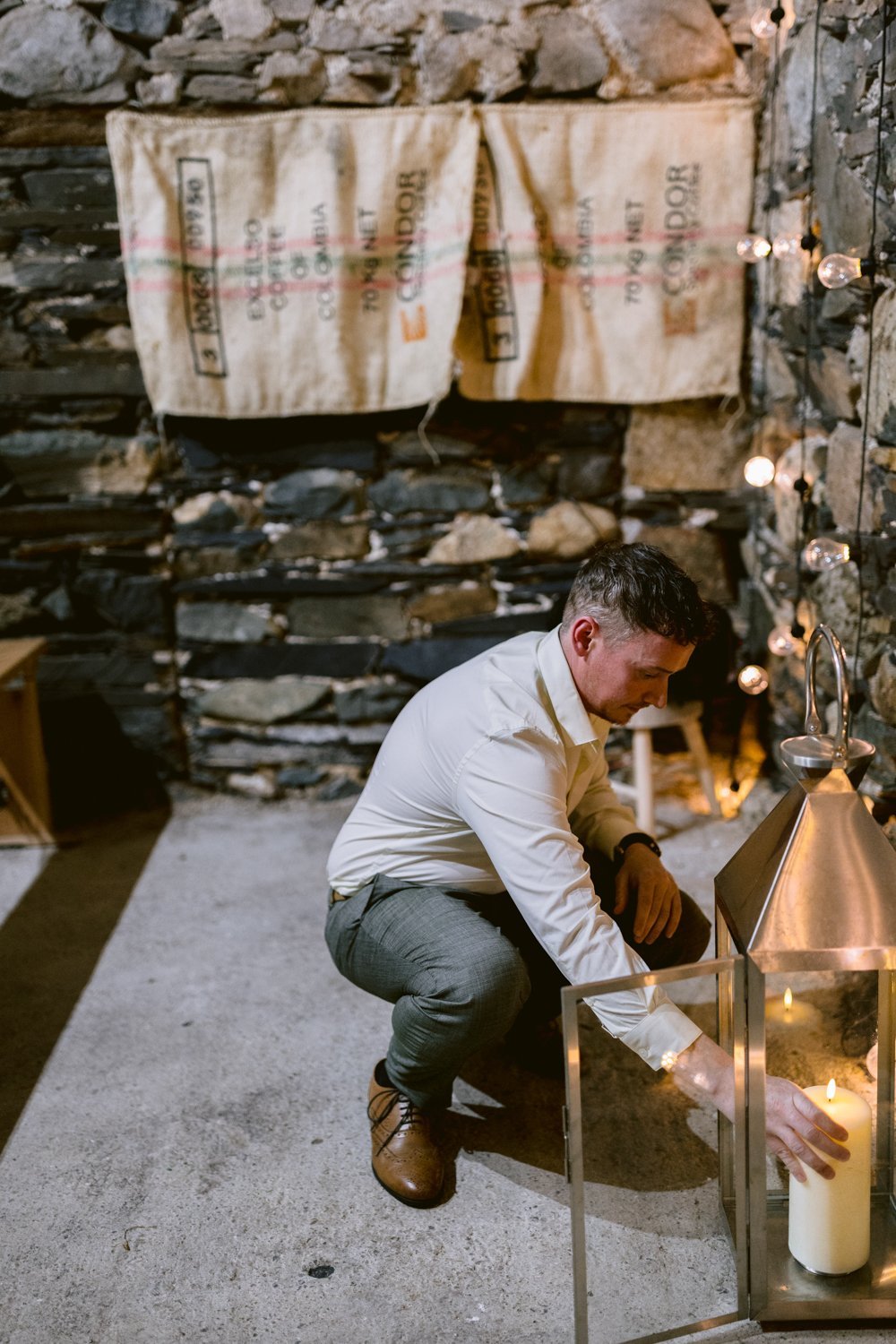 Man in a white shirt kneels to light a candle in a large lantern. Stone walls with burlap sacks and string lights are in the background.