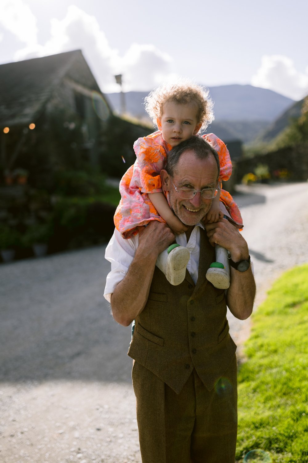 An older man carries a child on his shoulders outdoors, with a house and mountains in the background.