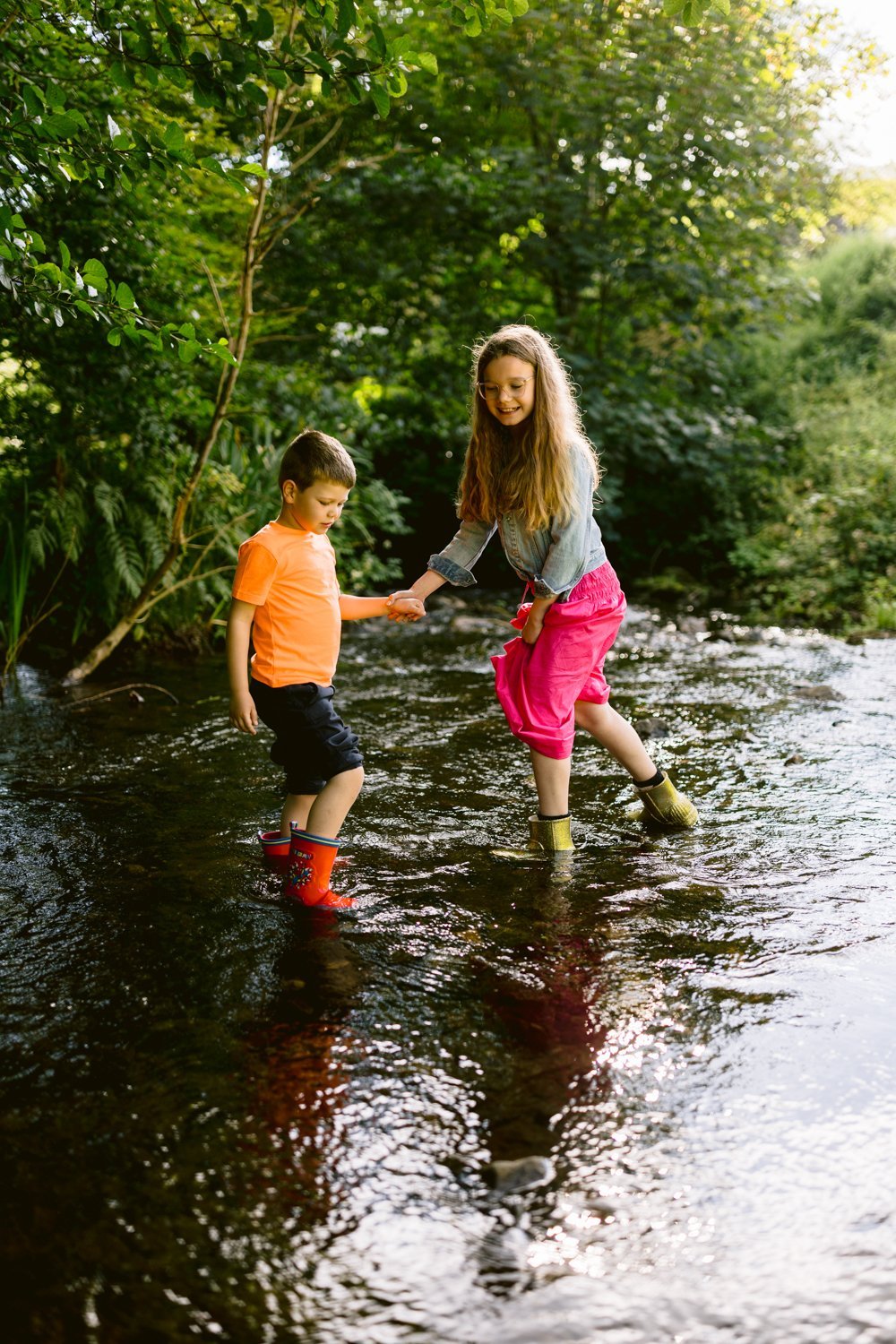 Two children wade through a shallow stream, holding hands. The boy wears an orange shirt and red boots, the girl a gray top and pink pants. Lush greenery surrounds them.