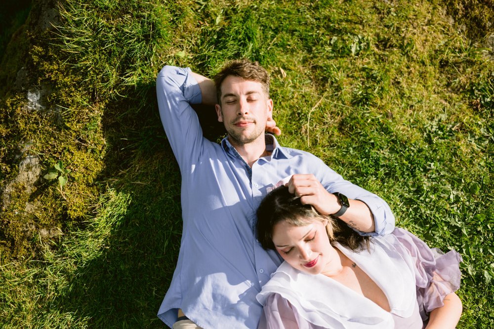 A man and a woman relax on green grass; he is reclining with hands behind head, and she rests her head on his shoulder, eyes closed.