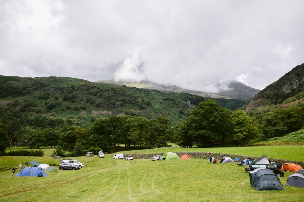 A grassy campsite with several tents and parked cars, surrounded by lush green hills and cloudy skies.