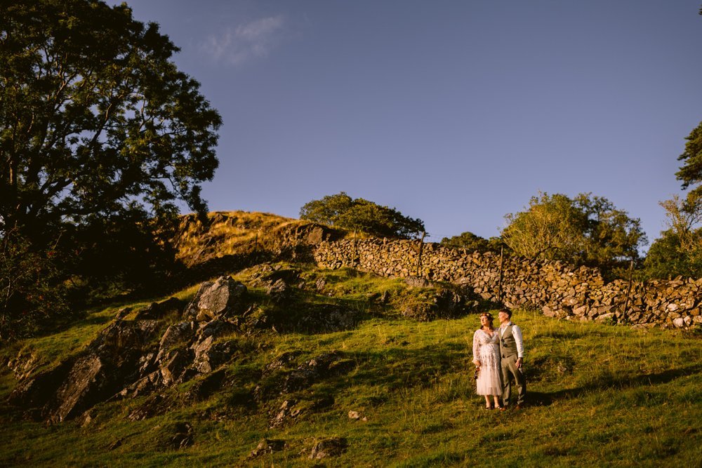 A couple stands on a grassy hill with a stone wall and trees in the background under a clear blue sky.