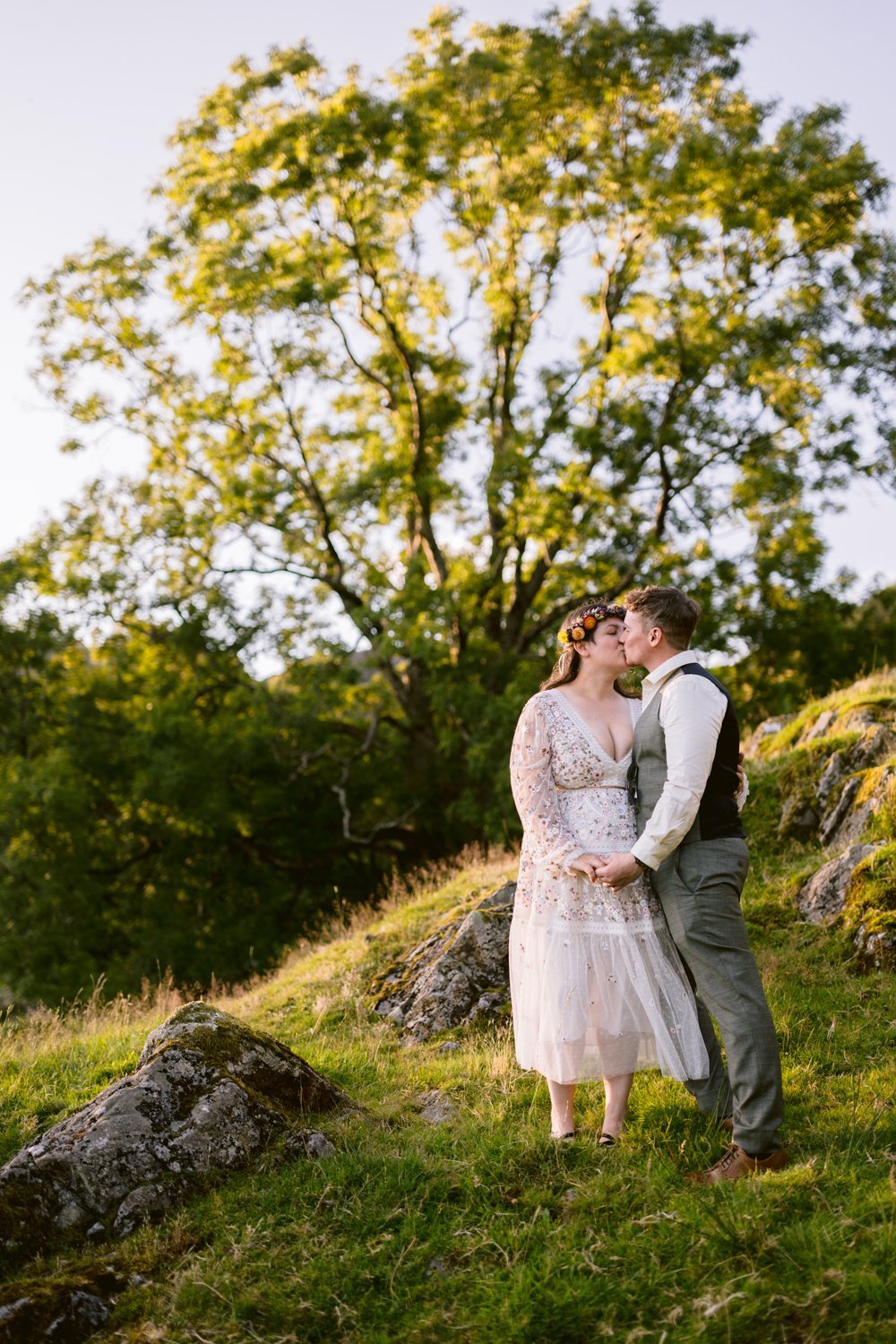A couple stands on a grassy hill, with a large tree in the background. The man kisses the woman's forehead as they hold hands.