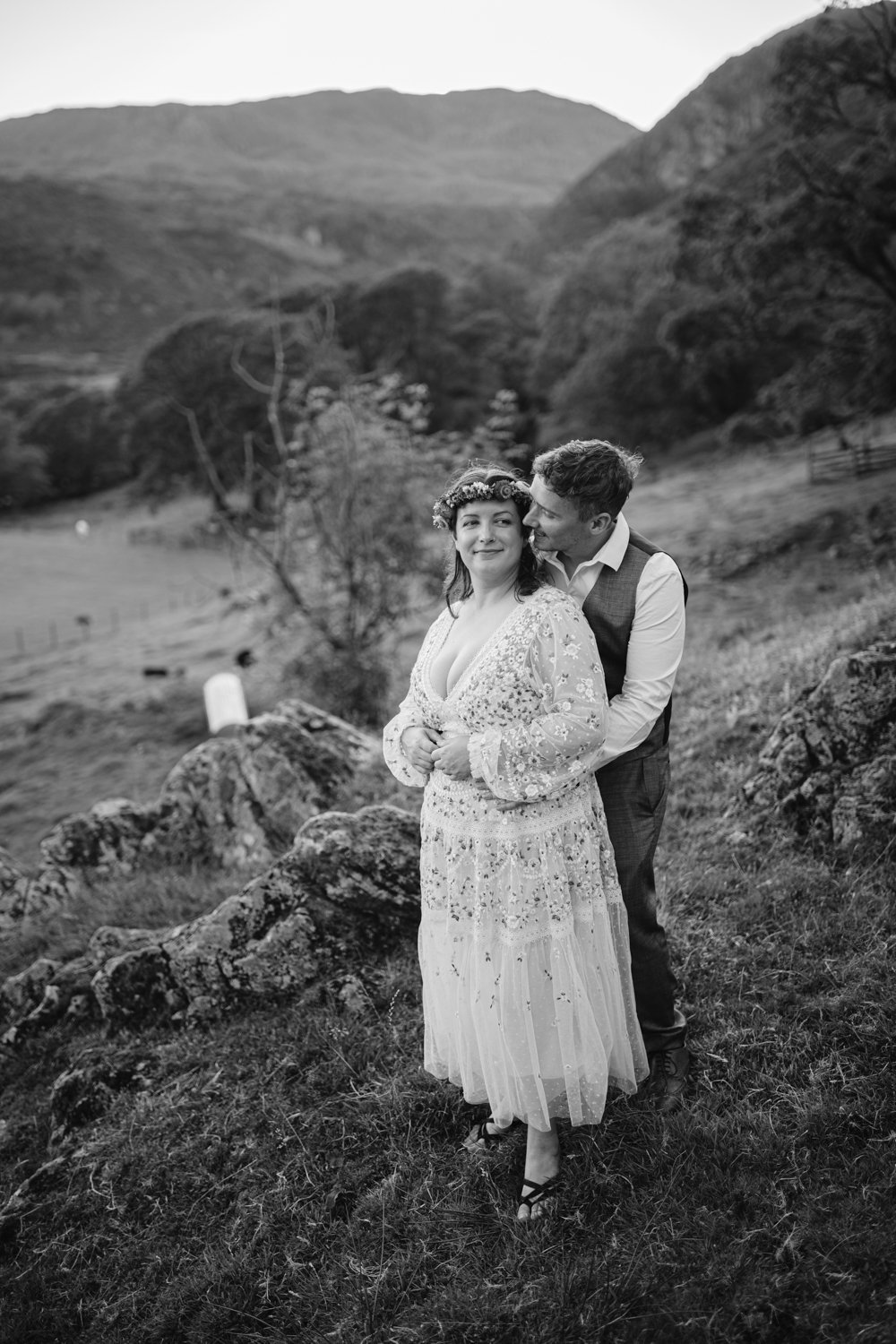 A couple in formal attire stands on a grassy hillside, with the man embracing the woman from behind. Trees and hills are visible in the background. The image is in black and white.