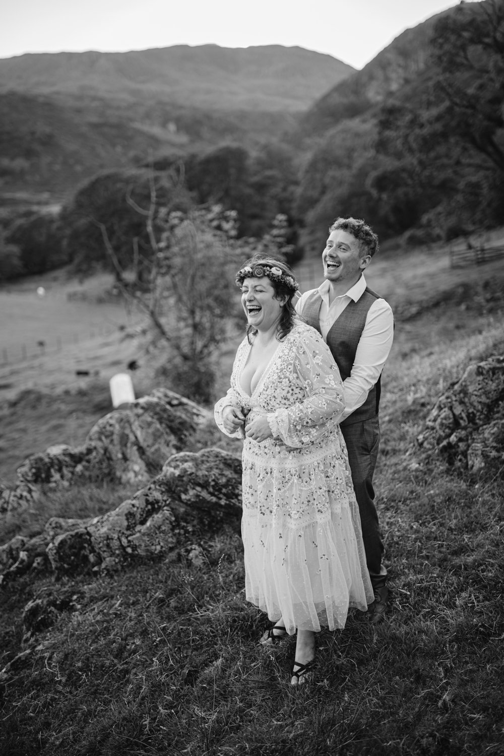 A couple laughing together on a grassy hillside, with mountains and trees in the background. They are dressed in wedding attire. Black and white image.