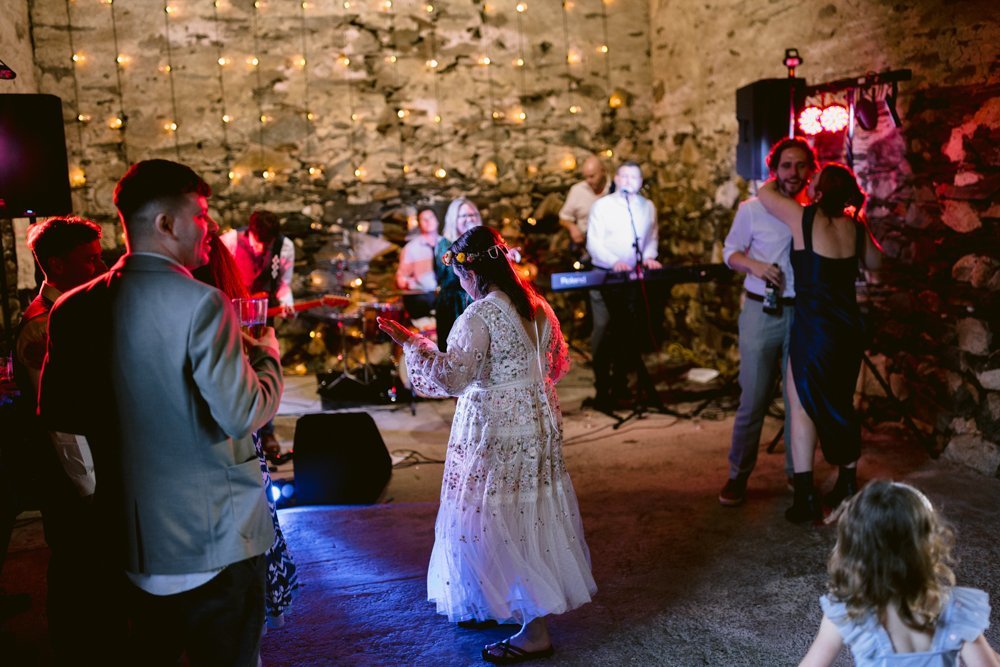 A group of people dance and socialize at a party in a rustic venue with stone walls and string lights.