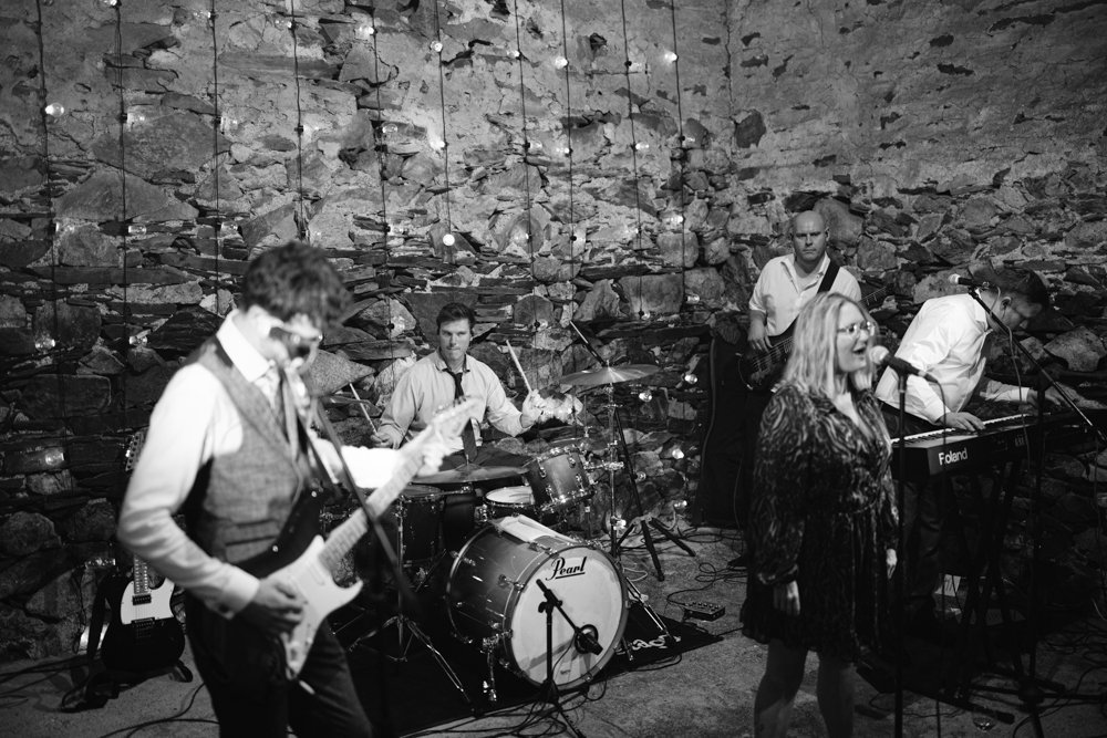 A band performs in front of a rustic stone wall, featuring a guitarist, drummer, keyboardist, and a female singer. Strings of lights hang overhead.
