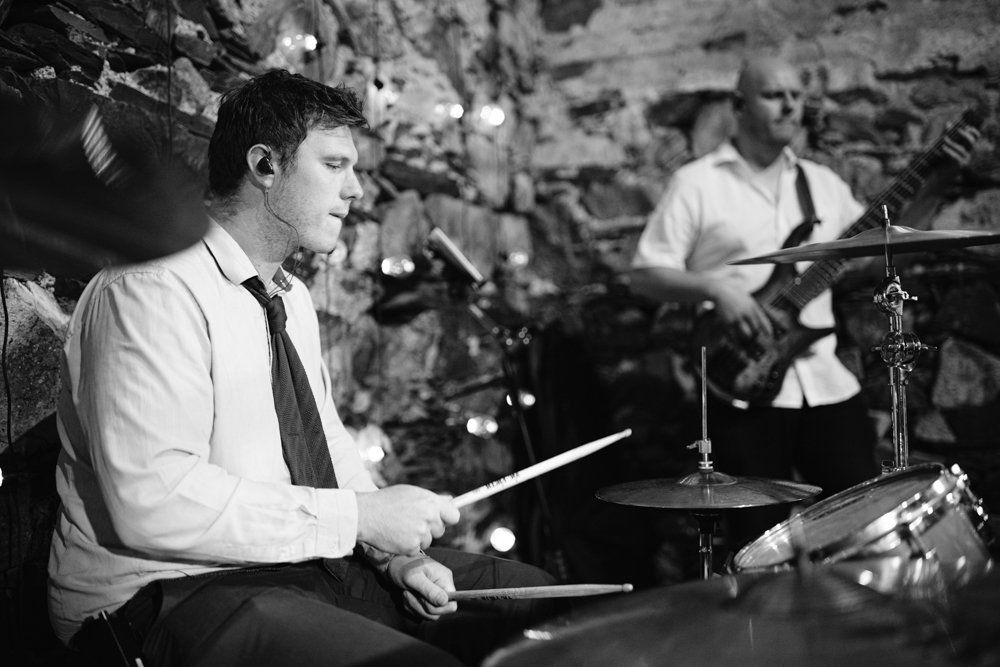 Drummer and bassist performing in a rustic setting with stone walls and string lights. Drummer focused on playing, while bassist stands behind. Black and white image.
