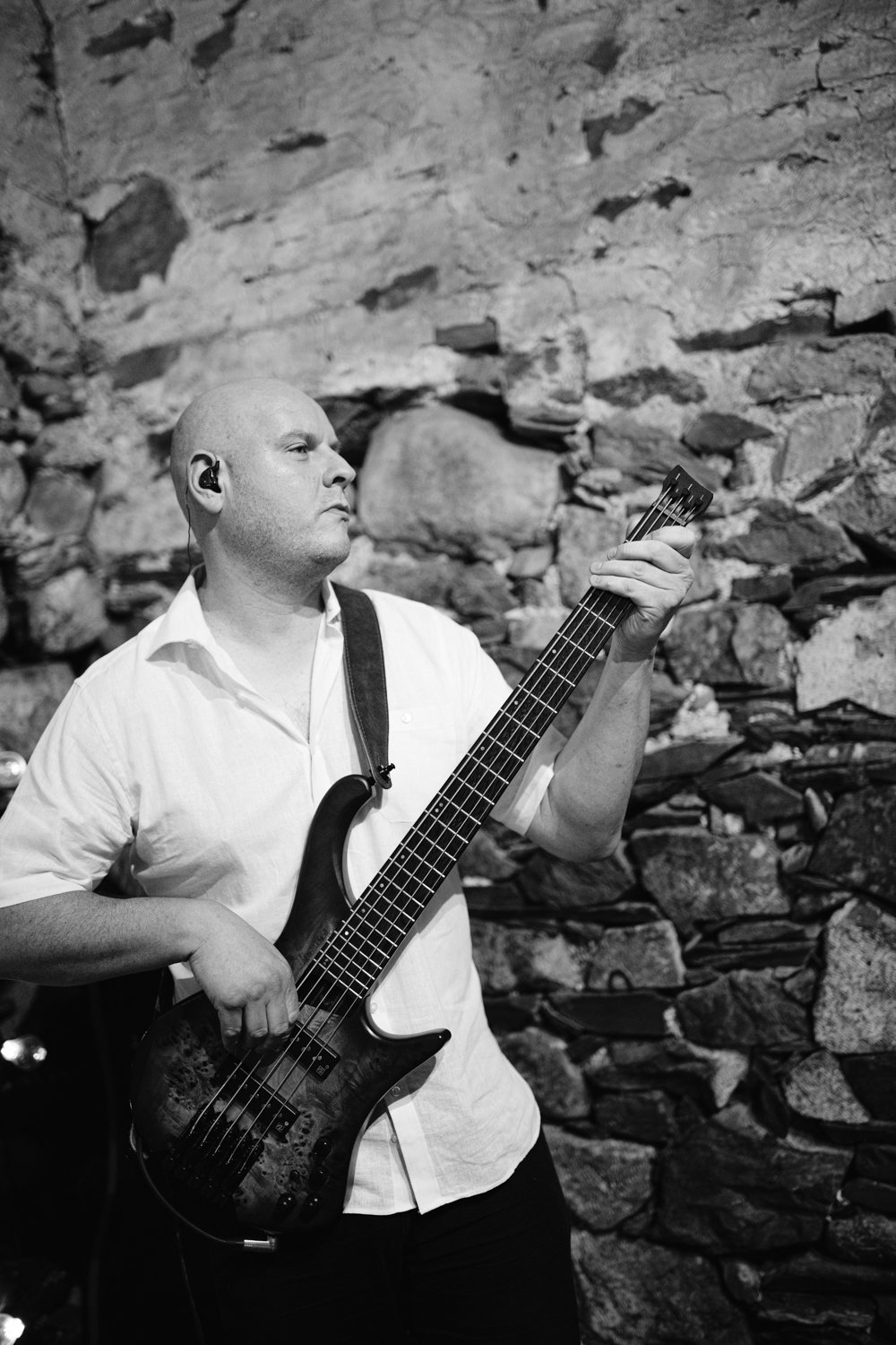 Man playing bass guitar in front of a stone wall, wearing a white shirt and in-ear monitors. Black and white photo.