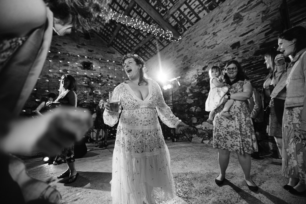 Woman in a white dress dances joyfully in a rustic venue. Surrounding people watch, one holding a child. String lights hang from the ceiling. Black and white photo.