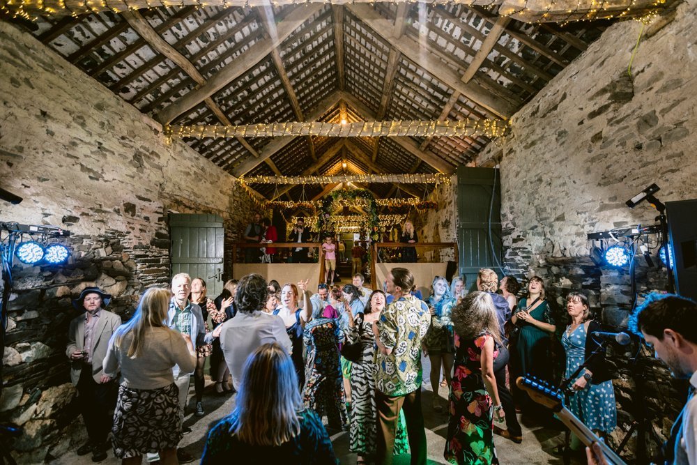 A group of people dance under string lights in a rustic stone barn with exposed beams.