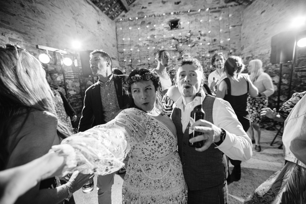 A couple dancing and smiling in a lively party setting, with others dancing in the background. The man holds a drink, and the woman extends her arm towards the camera.