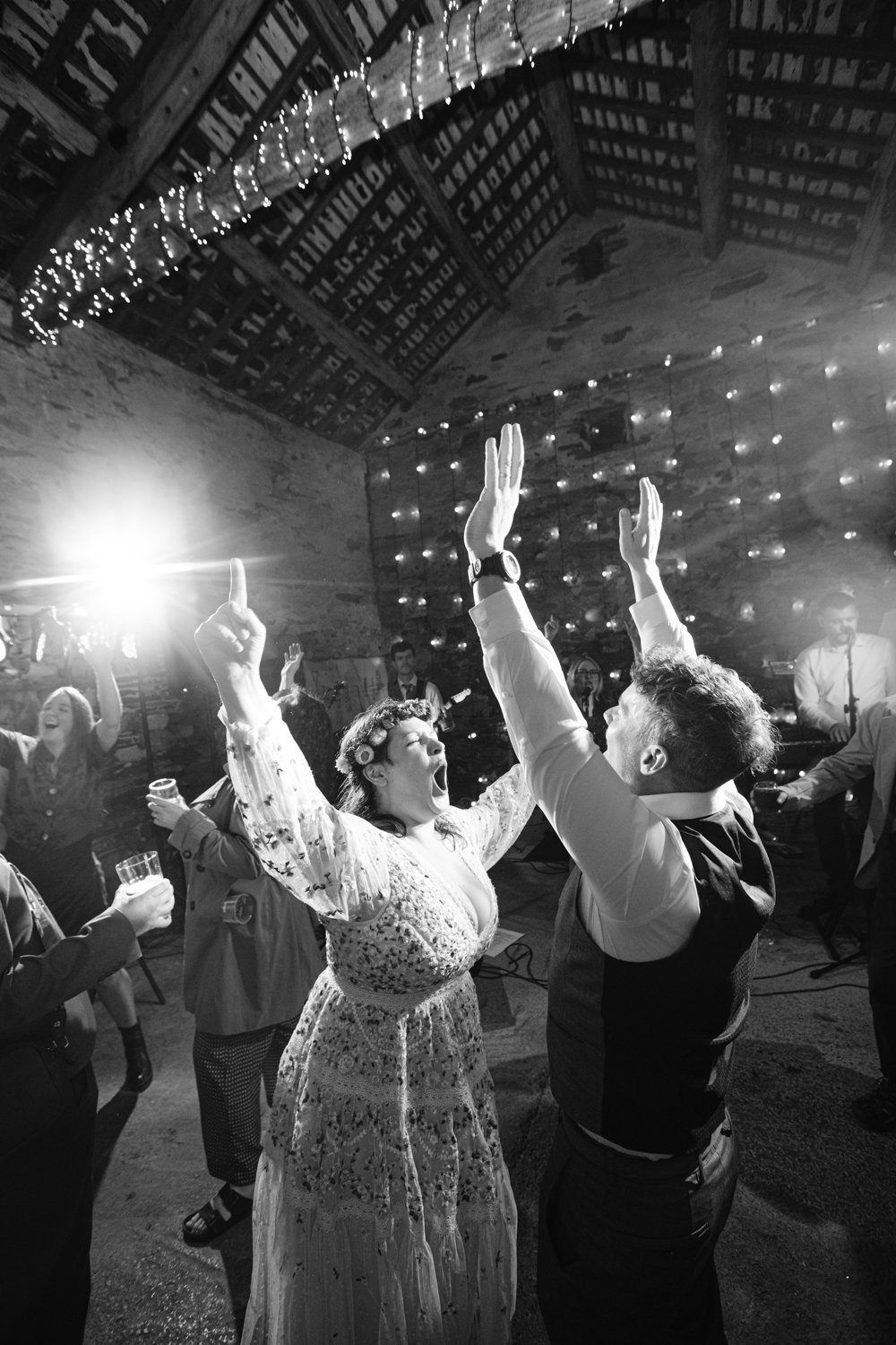 People dancing energetically at a celebration in a rustic venue with string lights and a spotlight.