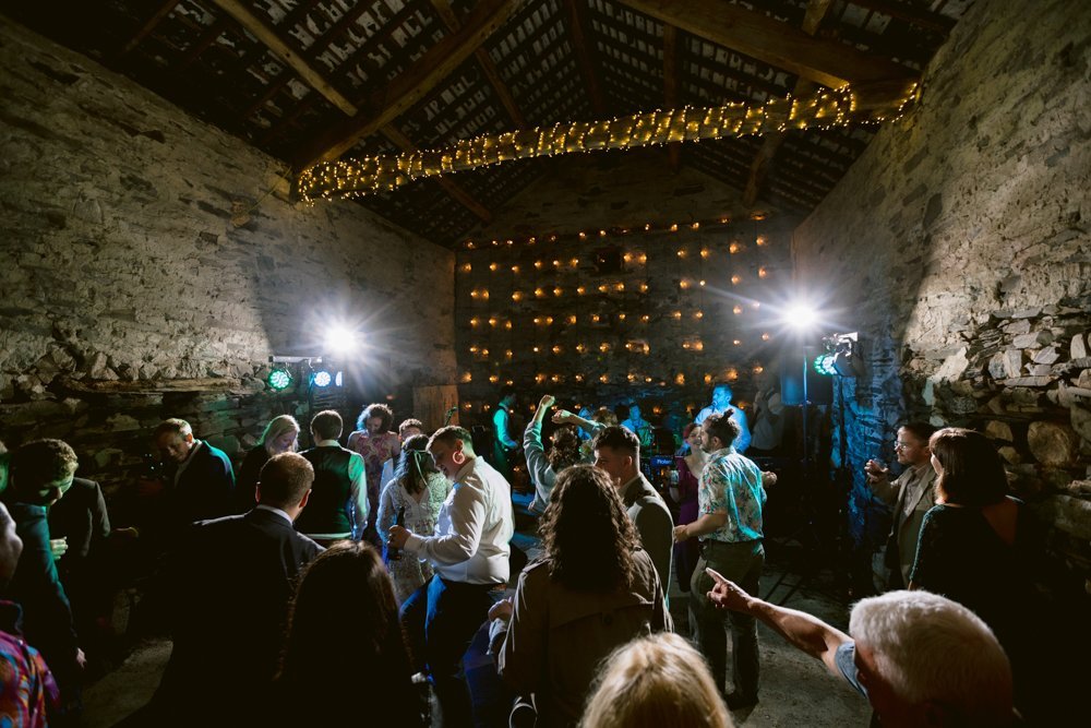 A group of people dance in a rustic stone building. String lights are hung on the ceiling and walls. Two bright lights illuminate the scene, creating a lively atmosphere.