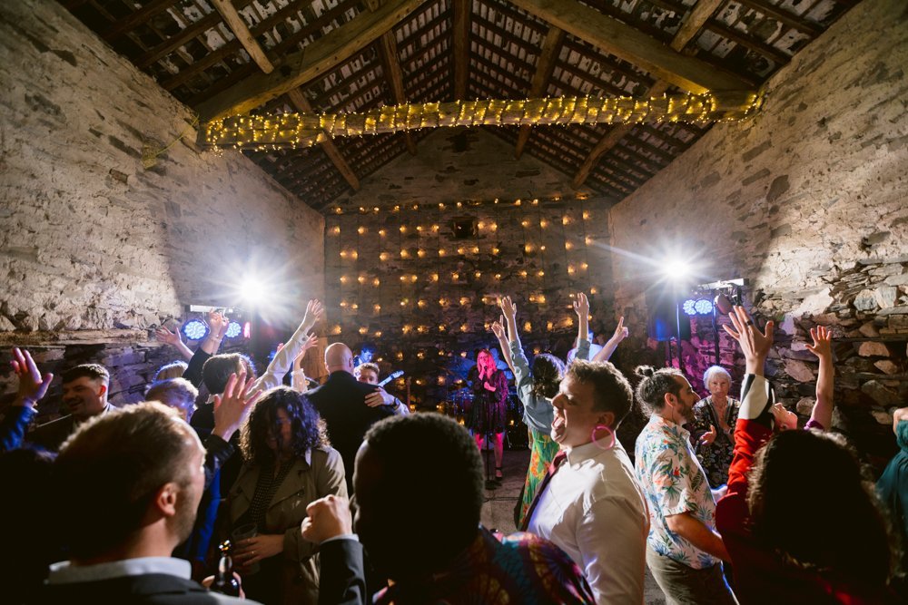 People dancing in a rustic barn decorated with string lights. The room is filled with music and vibrant lighting creating a lively atmosphere.