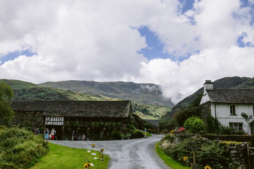 A rural scene with a road leading to stone buildings, surrounded by green hills and a cloudy sky. People gather near a barn-like structure.