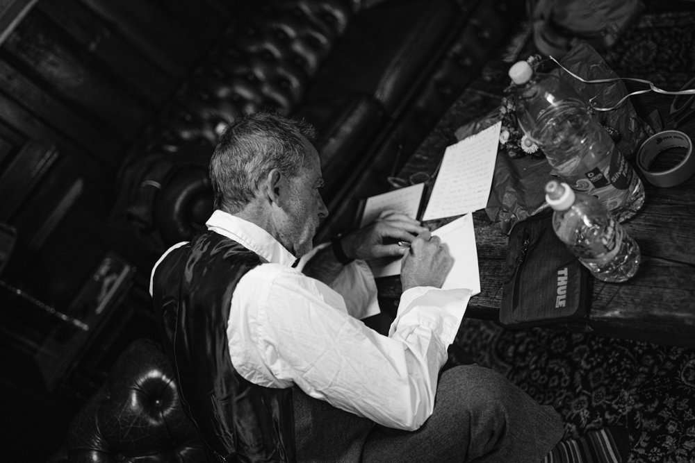 A man in a vest and shirt is writing on a piece of paper at a wooden table. Bottles and other items are scattered around. The setting appears to be a cozy room with leather furniture.