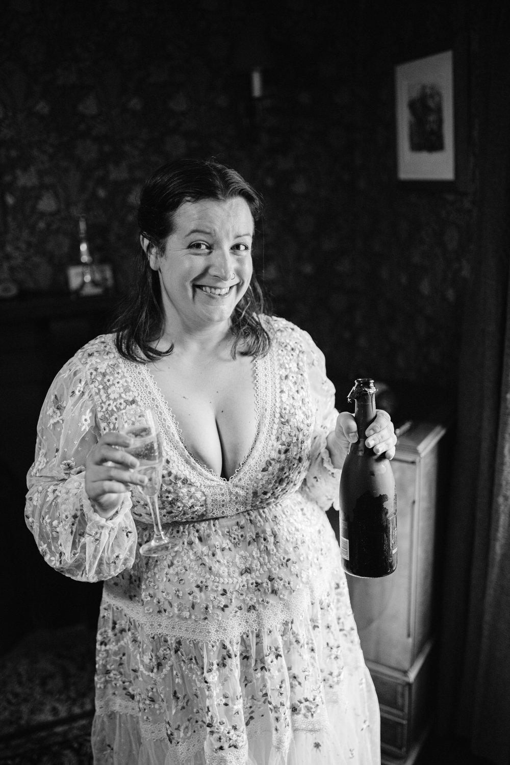 A woman in a floral dress holds a wine glass and a bottle, smiling at the camera, in a dimly lit room with patterned wallpaper. Black and white photo.