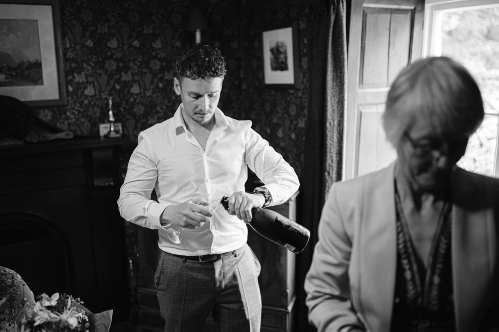 A man in a white shirt opens a bottle, standing near a fireplace. A person in a blazer is in the foreground. The setting appears to be indoors with framed pictures on the walls.