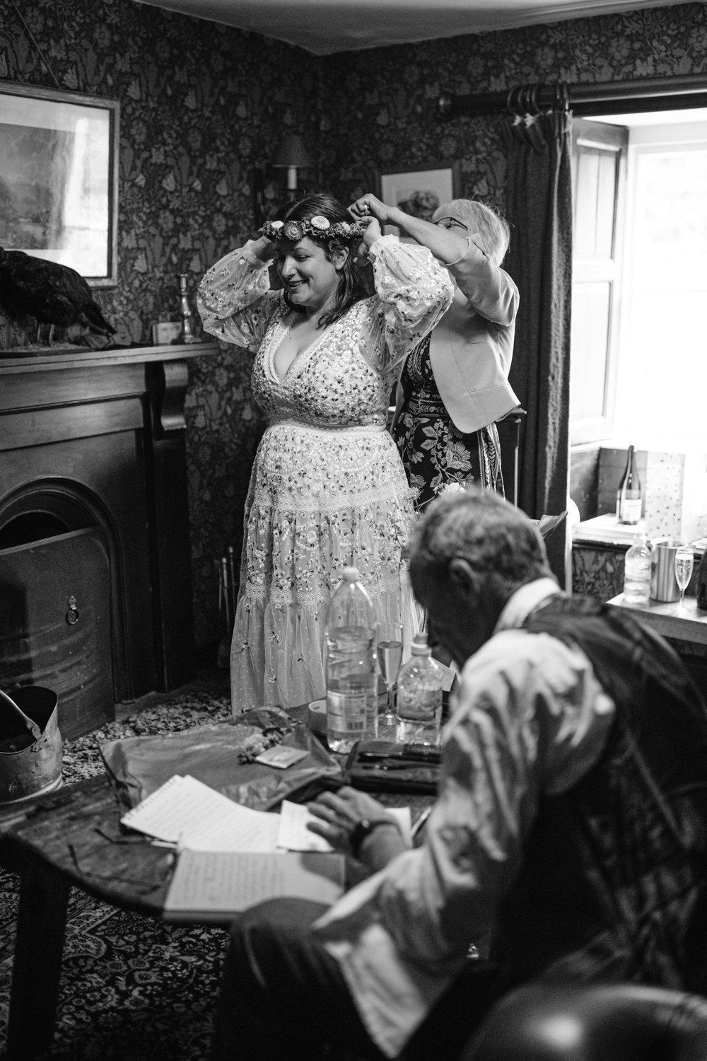 A woman in a floral dress adjusts a flower crown with assistance while another person sits at a table writing in a vintage-style room.