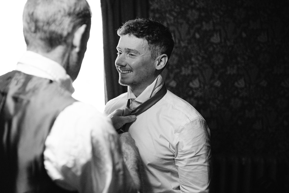 A person helps another person adjust their necktie in a dimly lit room. Both are dressed in formal attire.