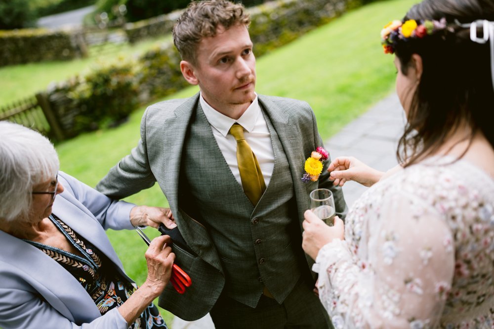 A man in a suit is assisted by two women, one adjusting his boutonniere and the other holding a pair of scissors. They're outdoors with grass and stone walls in the background.
