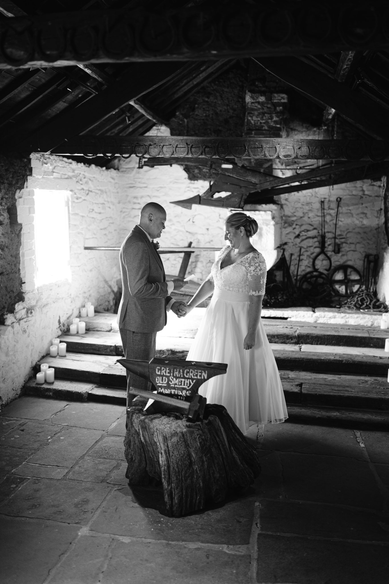 A couple holds hands during a wedding ceremony in a rustic, dimly lit space with candles and stone walls. A wooden sign with the words "Gretna Green Old Smithy" is in the foreground.