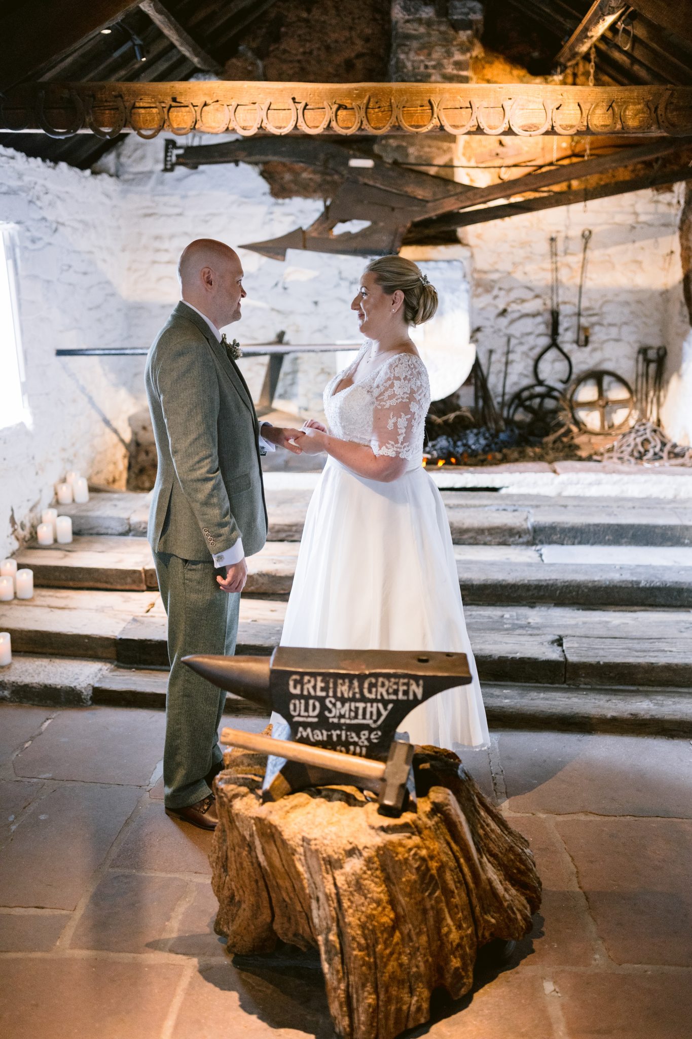 A couple stands facing each other in a rustic setting with a stone backdrop and an anvil labeled "Gretna Green Old Smithy Marriage.