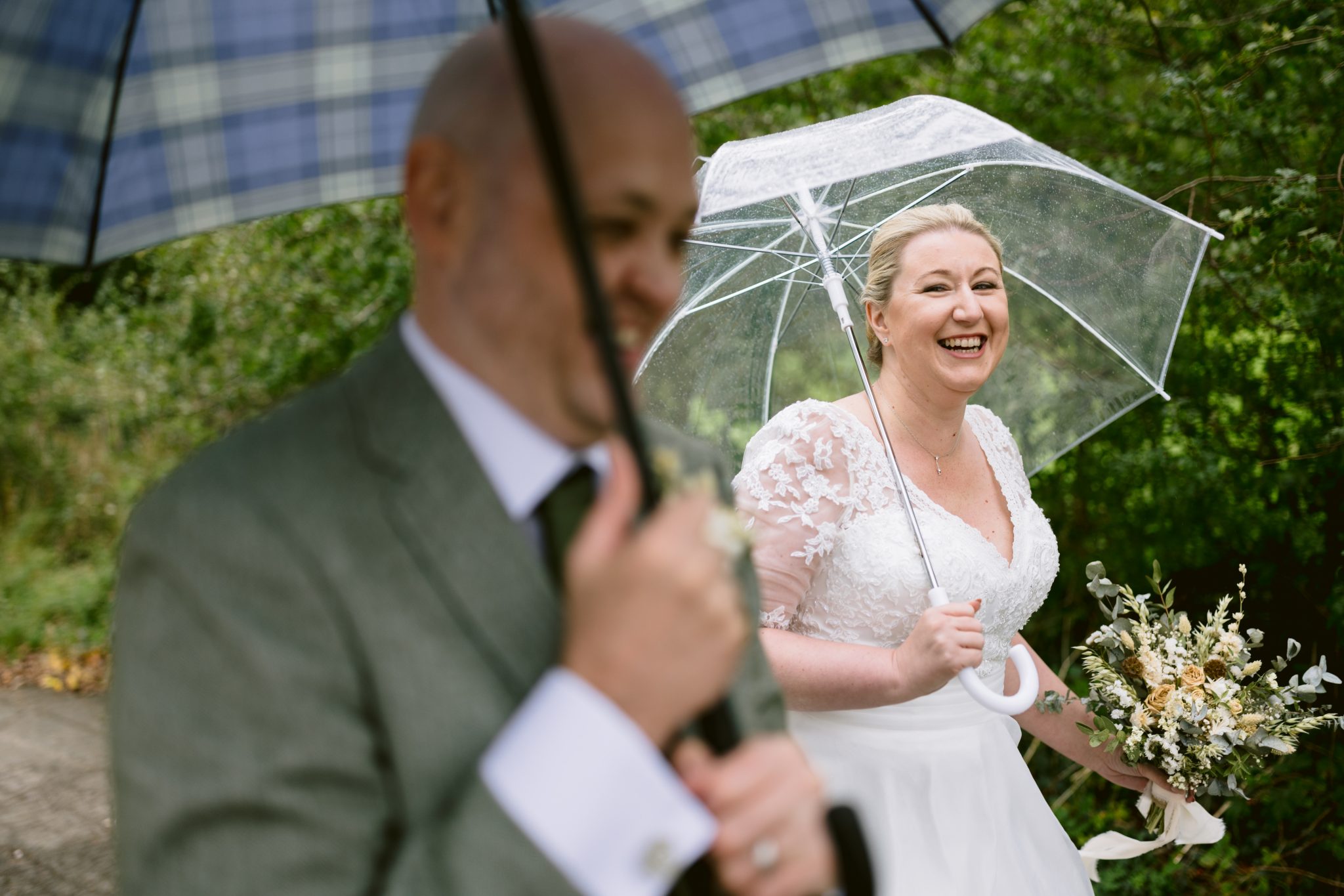 A smiling bride in a white dress holds a bouquet and a transparent umbrella. A suited man with a plaid umbrella is in the foreground. Green foliage is in the background.